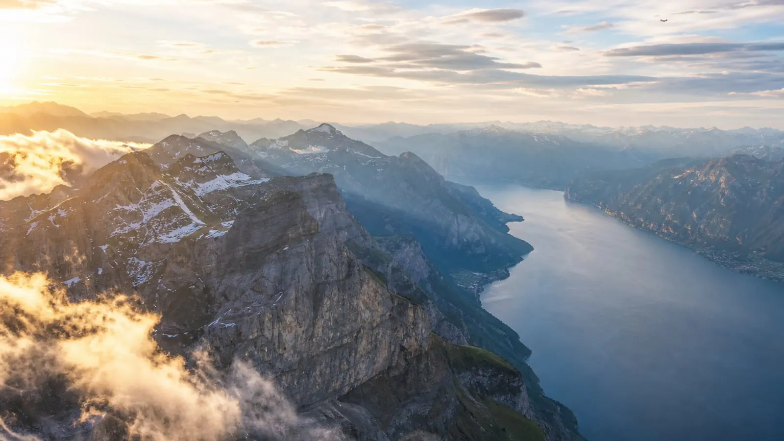 Dramatic aerial view over European mountains and lake at golden hour with a distant aircraft silhouette
